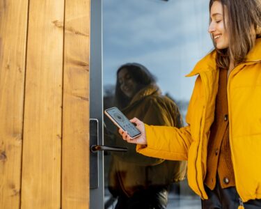 A woman in a yellow puffer coat uses her phone to unlock a smart lock on a wood-sided home with a large glass front door.