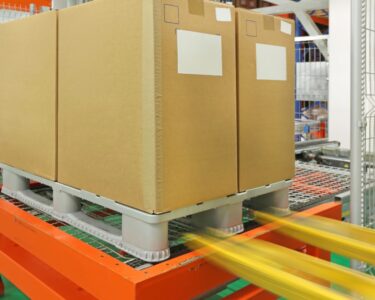 A plastic pallet with tall cardboard boxes being placed by a forklift onto a robotic palletizers in a warehouse.