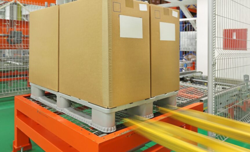 A plastic pallet with tall cardboard boxes being placed by a forklift onto a robotic palletizers in a warehouse.