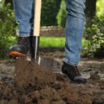 A close-up of a person wearing black boots and blue jeans stepping on a spade to push the shovel into dirt outdoors.