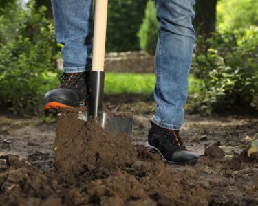 A close-up of a person wearing black boots and blue jeans stepping on a spade to push the shovel into dirt outdoors.