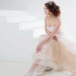 A woman in a light-colored dress sits on white steps, with flowing fabric spread around her in a bright, minimal setting.