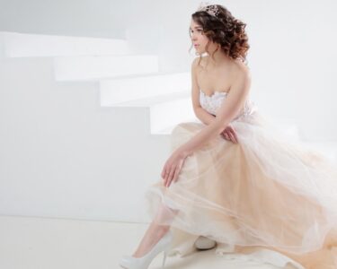 A woman in a light-colored dress sits on white steps, with flowing fabric spread around her in a bright, minimal setting.
