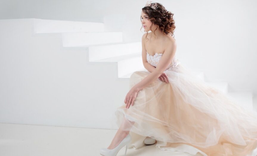 A woman in a light-colored dress sits on white steps, with flowing fabric spread around her in a bright, minimal setting.