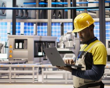 A man in a yellow hard hat and overalls looks at a laptop in an industrial facility with a conveyor belt in the background.