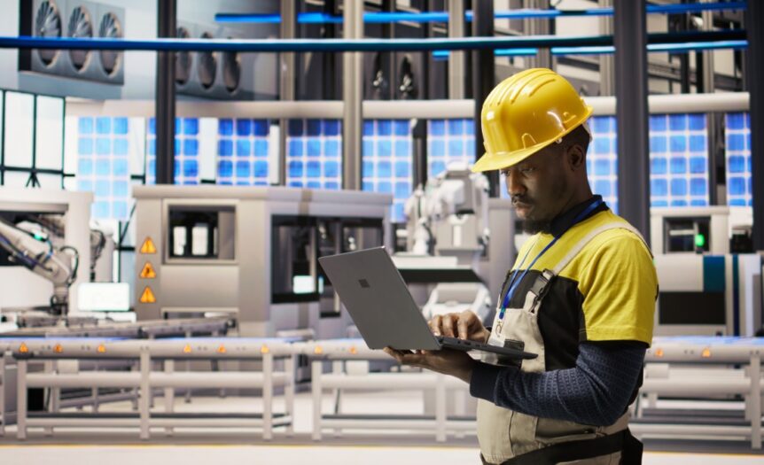 A man in a yellow hard hat and overalls looks at a laptop in an industrial facility with a conveyor belt in the background.