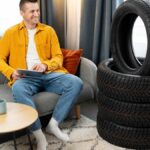 A man in a yellow shirt and jeans sits on a couch at home using a tablet with a stack of new car tires next to him.