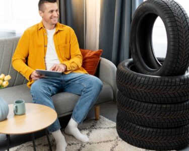 A man in a yellow shirt and jeans sits on a couch at home using a tablet with a stack of new car tires next to him.