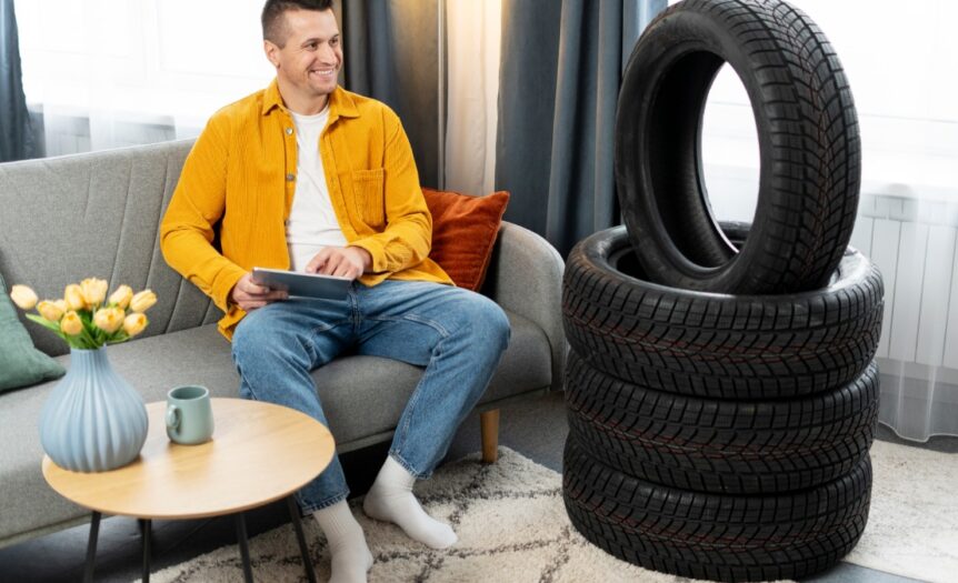 A man in a yellow shirt and jeans sits on a couch at home using a tablet with a stack of new car tires next to him.