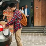 A son placing boxes in the trunk of his car with his parents looking at him from the porch of their house.