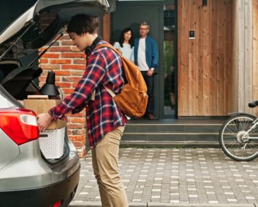 A son placing boxes in the trunk of his car with his parents looking at him from the porch of their house.