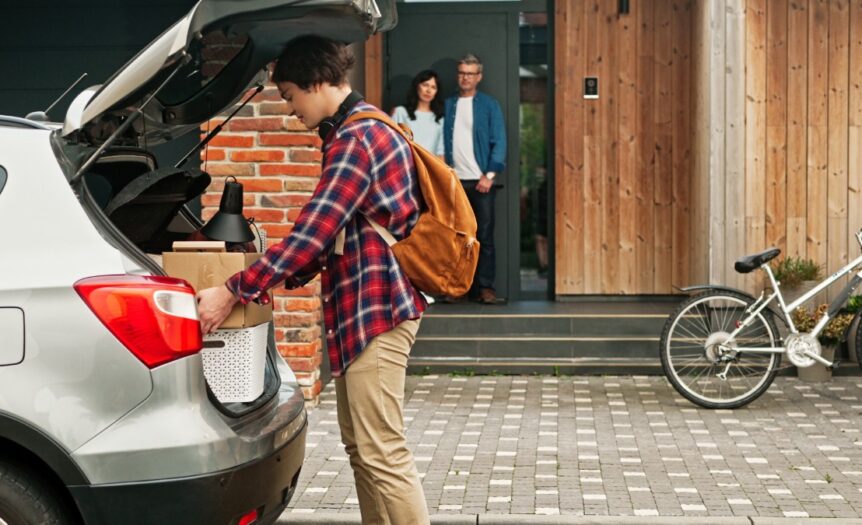 A son placing boxes in the trunk of his car with his parents looking at him from the porch of their house.