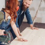 A woman holds a power saw while a man steadies pale wood planks in a room under renovation, with a ladder in back.