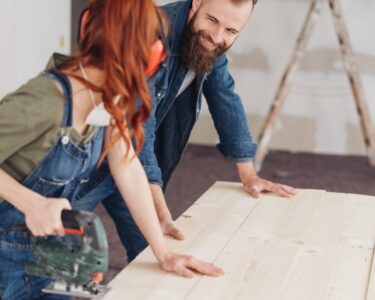 A woman holds a power saw while a man steadies pale wood planks in a room under renovation, with a ladder in back.