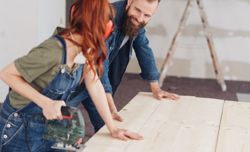 A woman holds a power saw while a man steadies pale wood planks in a room under renovation, with a ladder in back.
