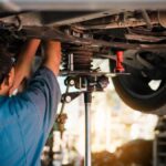 A man with glasses and a blue shirt facing away from the viewer working on the undercarriage of a car on a lift.