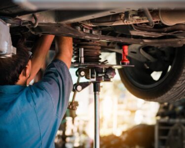 A man with glasses and a blue shirt facing away from the viewer working on the undercarriage of a car on a lift.