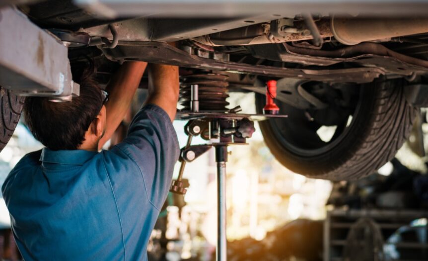 A man with glasses and a blue shirt facing away from the viewer working on the undercarriage of a car on a lift.