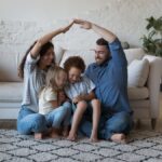 A family with a mother, father, and two children are sitting on the floor of a living room. There is a couch behind them.