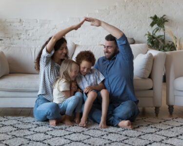 A family with a mother, father, and two children are sitting on the floor of a living room. There is a couch behind them.