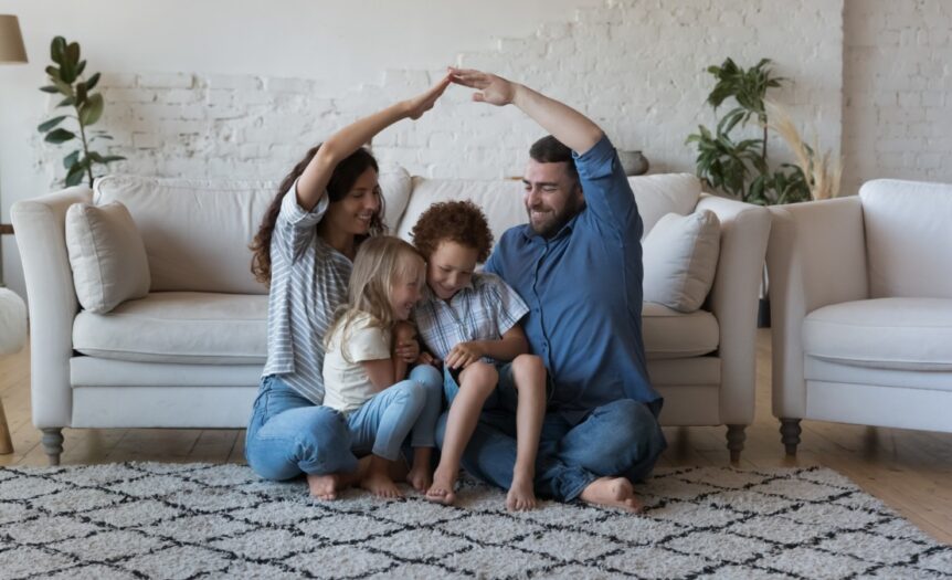 A family with a mother, father, and two children are sitting on the floor of a living room. There is a couch behind them.