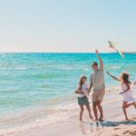 Parents and children running on sandy beach flying colorful kite beside ocean, with sunlight reflecting on water.