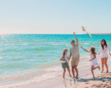 Parents and children running on sandy beach flying colorful kite beside ocean, with sunlight reflecting on water.