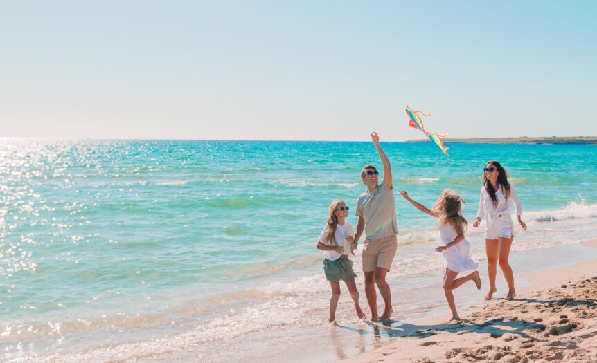 Parents and children running on sandy beach flying colorful kite beside ocean, with sunlight reflecting on water.