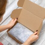 A pair of hands places bubble wrap into an open cardboard box, with a pair of scissors lying nearby on a light surface.