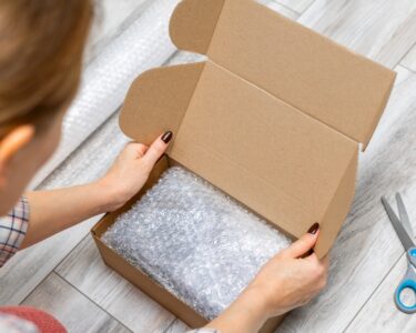 A pair of hands places bubble wrap into an open cardboard box, with a pair of scissors lying nearby on a light surface.