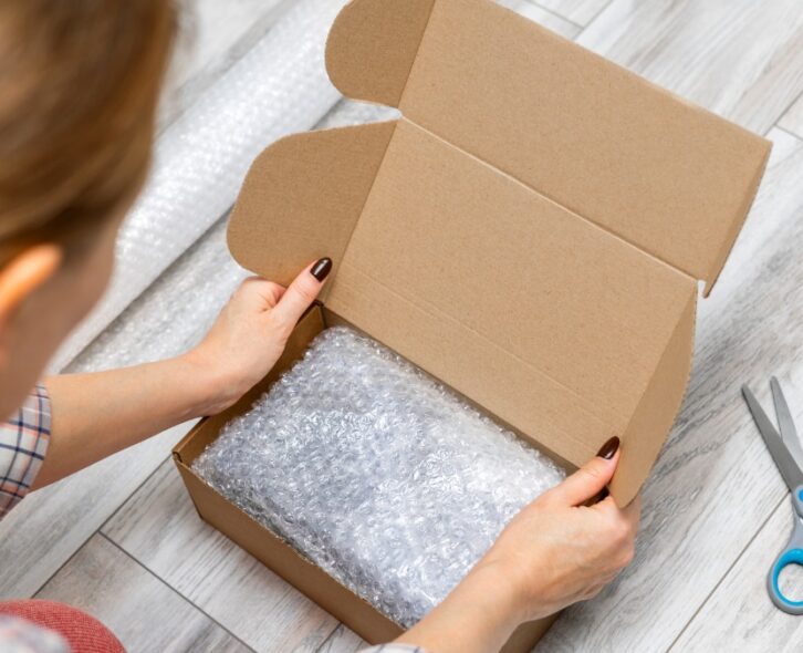 A pair of hands places bubble wrap into an open cardboard box, with a pair of scissors lying nearby on a light surface.