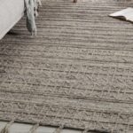 A large gray rug lies on the floor of a home next to a gray couch and a small brown stool. A window sits nearby.