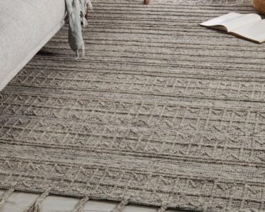 A large gray rug lies on the floor of a home next to a gray couch and a small brown stool. A window sits nearby.
