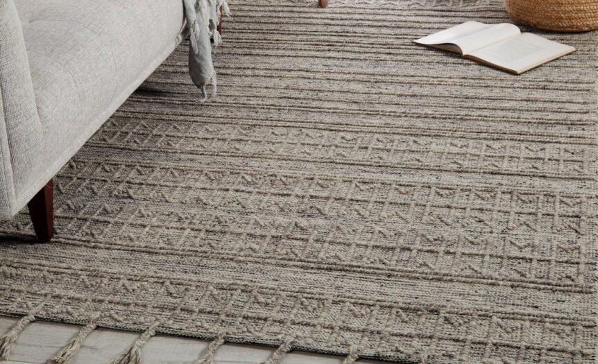 A large gray rug lies on the floor of a home next to a gray couch and a small brown stool. A window sits nearby.