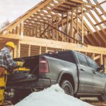A man in a yellow hard hat is standing next to a pickup truck which is parked in front of a house that us under construction.