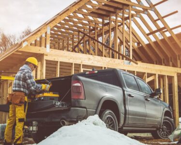 A man in a yellow hard hat is standing next to a pickup truck which is parked in front of a house that us under construction.