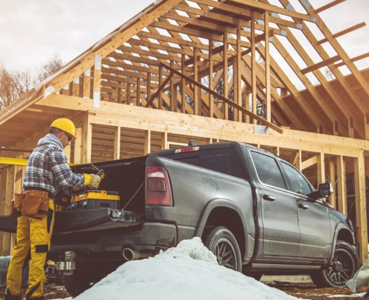 A man in a yellow hard hat is standing next to a pickup truck which is parked in front of a house that us under construction.