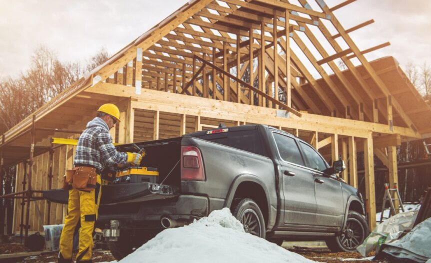 A man in a yellow hard hat is standing next to a pickup truck which is parked in front of a house that us under construction.