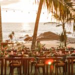 A table with chairs is set up on a sandy beach. On the table is an assortment of plates and flowers.