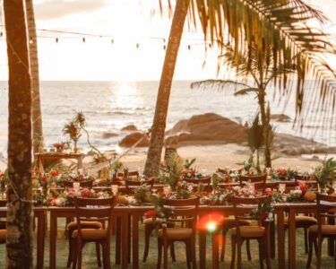 A table with chairs is set up on a sandy beach. On the table is an assortment of plates and flowers.