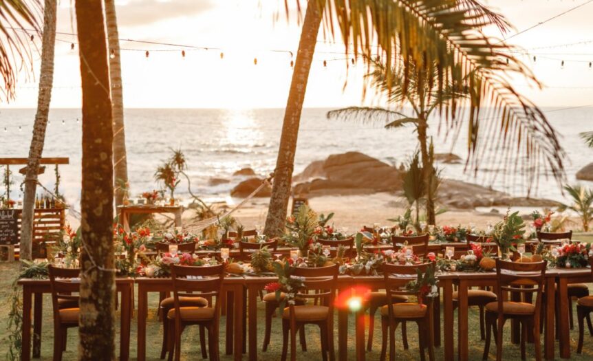 A table with chairs is set up on a sandy beach. On the table is an assortment of plates and flowers.