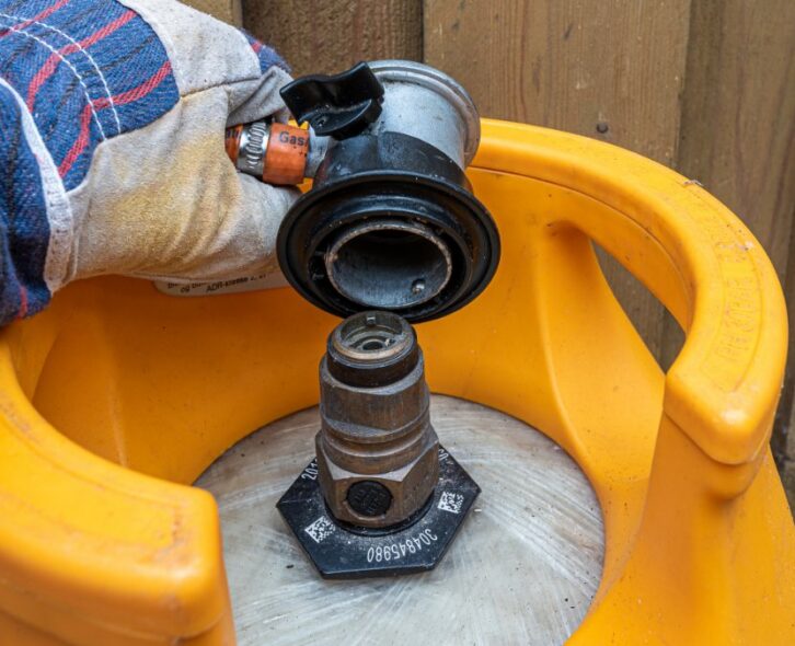 A close-up view shows a person wearing a gray glove placing a gas regulator on a gas cylinder that sits outside.