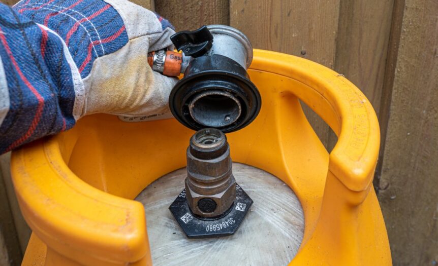 A close-up view shows a person wearing a gray glove placing a gas regulator on a gas cylinder that sits outside.
