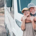 An older man and woman are standing in front of a park RV. The man is behind the woman, wrapping his arms around her.