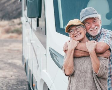 An older man and woman are standing in front of a park RV. The man is behind the woman, wrapping his arms around her.
