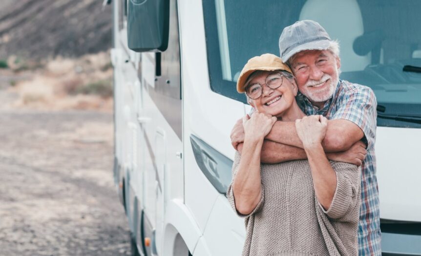An older man and woman are standing in front of a park RV. The man is behind the woman, wrapping his arms around her.