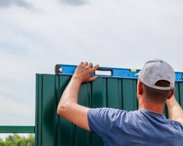 A person in a gray cap holding a blue level across the top of a green metal fence under a cloudy sky.