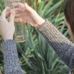 A woman's two hands holding a clear rain gauge with visible markings and water, surrounded by green garden foliage.