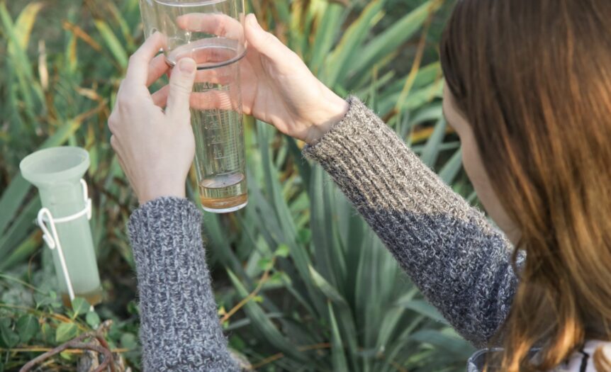 A woman's two hands holding a clear rain gauge with visible markings and water, surrounded by green garden foliage.
