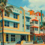 Palm trees line pastel Art Deco buildings along a sunny street under a bright blue sky with cream and coral facades.
